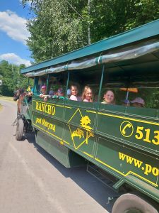 Dzieci siedzą w zielonym autobusie bez okien. Na autobusie widnieje napis "RANCHO pod Olszyną" oraz logo rancha.