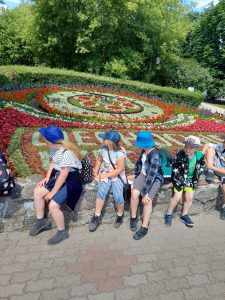 Uczniowie siedzą na murku i odwracają się za siebie. Patrzą na ułożony z kwiatów napis "CIECHOCINEK".