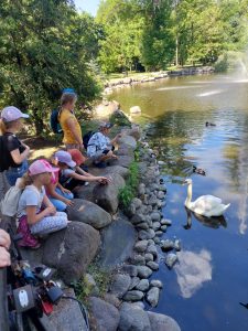 Uczniowie nachylają się nad stawem i karmią łabędzie, który do nich podpłynął.