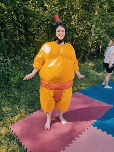 Uczennica w nadmuchanym, pomarańczowym stroju sumoki stoi na kolorowej macie. Dziewczynka uśmiecha się.