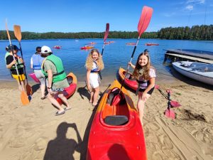 Dwie uczennice pozują do wspólnego zdjęcia przy kajaku leżącym na plaży. Dziewczynki w dłoniach trzymają wiosła. W tle widać innych uczniów stojących z wiosłami oraz kajaki pływające po jeziorze.