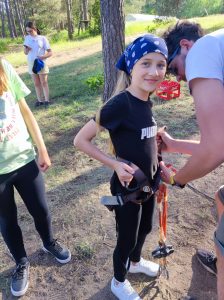 Uczennica podczas zapinania liny do uprzęży. Dziewczynka uśmiecha się. Przy niej stoi Pan mocujący linę.