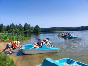 Uczniowie siedzą w rowerach wodnych. Dwie uczennice odpychają jeden z rowerów od brzegu.