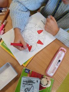 Zbliżenie na szkolna ławkę, na której leży wykonywana przez uczennicę mandala. Dziewczynka maluje jej elementy czerwonym pisakiem.