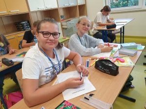 Dwie uczennice uśmiechają się siedząc przy szkolnej ławce. Na stoliku przed nimi leżą kartki papieru i przybory szkolne.