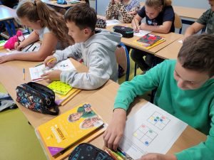 Uczniowie klasy czwartej siedzą przy wspólnym stole i wykonują zadania z okazji Dnia Kropki. Przed dziećmi leżą kartki z sudoku otworzonym z kolorowych kropek.