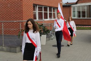 Troje uczniów wprowadza poczet flagowy na apel. Wszyscy mają przepasane przez ramię biało czerwone szarfy. Uczeń idący po środku niesie flagę Polski.