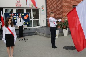 Na pierwszym planie stoją w odstępie od siebie uczeń i uczennica przepasani biało czerwonymi szarfami. Uczeń trzyma flagę Polski opuszczoną do przodu. W tle widać stojących na baczność dwoje uczniów prowadzących apel z okazji rozpoczęcia roku szkolnego.