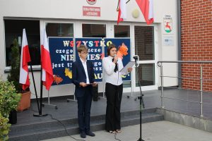 Dwoje uczniów prowadzących apel stoi obok siebie przed budynkiem szkoły. Za nimi ustawiona jest tablica z kolorowym napisem.