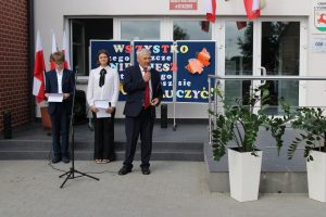 Przed wejściem do szkoły stoi dwoje uczniów prowadzących apel a obok nich Pan wójt trzymający mikrofon. Za uczniami stoi tablica z kolorowym napisem ozdobiona pomarańczowym motylem.