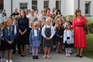 Uczniowie stoją w biało czarnych stroją na szkolnym boisku przed wejściem do szkoły. Po prawej stronie stoi ubrana w czerwoną sukienkę nauczycielka.