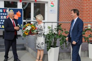 Pani Dyrektor otrzymuje bukiet kwiatów z rak Pana wójta. Obok stoi komisarz do spraw Oświaty. Zdjęcie wykonane przed budynkiem szkoły.