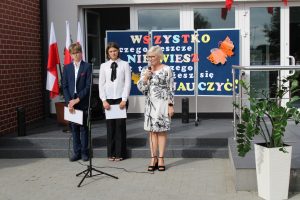 Przed wejściem do szkoły stoi dwoje uczniów prowadzących apel a obok nich Pani Dyrektor trzymająca mikrofon. Za uczniami stoi tablica z kolorowym napisem ozdobiona pomarańczowym motylem.