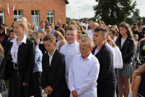 Uczniowie stoją w biało czarnych stroją na szkolnym boisku przed wejściem do szkoły.