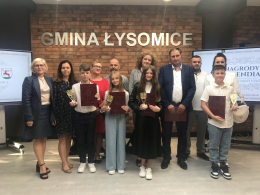 Zdjęcie grupowe wykonane na tle ściany z cegieł z umieszczonym centralnie napisem "GMINA ŁYSOMICE". Do zdjęcia pozują uczniowie naszej szkoły, którzy otrzymali stypendia wraz z rodzicami i Panią Dyrektor. Uczniowie trzymają w dłoniach bordowe teczki i otrzymane puchary.