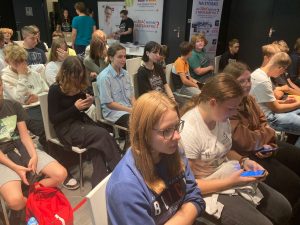 Uczniowie klasy ósmej siedzą na krzesełkach podczas spotkania motywacyjnego matma^2. Uczniowie spoglądają w swoje telefony.