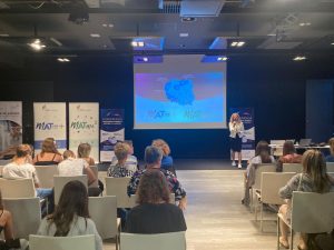 Zdjęcie wykonane podczas przemówienia jednej z organizatorek spotkania - przedstawicielki wydawnictwa elitmat. Pani stoi na tle rzutniki na którym wyświetla się mapa Polski z zaznaczonymi województwami biorącymi udział w projekcie. Obok ustawione są banery promujące projekty matma^2 i matura+.