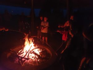 Uczniowie klasy 5 stoją przy ognisku i pieką kiełbaski. Zdjęcie ciemne z centralnie umieszczonym jasnym ogniskiem.