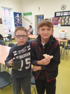 Dwóch uczniów klasy piątej pozuje do wspólnego zdjęcia stojąc na szkolnej stołówce. Jeden chłopiec trzyma w dłoni kubek a drugi tost.