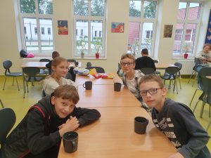 Czworo uczniów klasy piątej pochyla się nad długim stołem na szkolnej stołówce. Na stole stoja kubki z herbatą. Uczniowie spoglądaja w stronę aparatu.