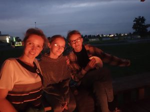 Dwie uczennice klasy piątej pozują do zdjęcia z nauczycielką. Zdjęcie wykonane nocą. Postacie oświetlone są światłem z ogniska.