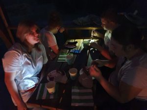 Uczniowie klasy piątej siedzą przy stoliku i jedzą kiełbaski. Zdjęcie wykonane nocą. Postacie są słabo widoczne.