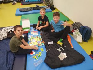 Trzech uczniów siedzi na materacach ułożonych na podłodze sali teatralnej. Pomiędzy nimi leży gra planszowa. Chłopcy uśmiechają się do obiektywu.