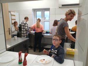 Zdjęcie wykonane podczas śniadania na szkolnej stołówce. Na pierwszym planie w oknie wydawczym siedzi uczeń jedzący tost. Za nim stoi nauczycielka spoglądająca na niego. W tle widać uczennice stojące przy stole plecami do zdjęcia.