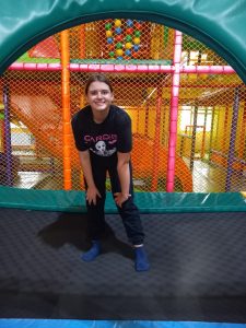 Uczennica klasy siódmej pozuje do zdjęcia stojąc na trampolinie. Dziewczynka lekko pochyla się do przodu opierając dłonie o kolana i szeroko się uśmiecha.