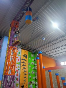 Uczennica klasy siódmej stoi na trampolinie zamocowanej pod sufitem. Dziewczynka zapięta jest w uprząż a na głowie ma kask.