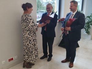 Wójt gminy Łysomice wraz z przewodniczącym Rady gminy wręczają nauczycielce stojącej bokiem do zdjęcia nagrodę. Wójt trzyma dyplom umieszczony w sztywnej oprawie a przewodniczący kwiaty.