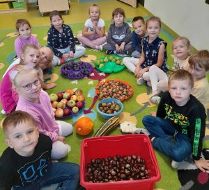 Uczniowie oddzialu przedszkolnego siedzą na dywanie. Między nimi rozłożone są w miskach i na talerzach dary jesieni - kasztany, orzechy, jabłka śliwki, gruszki oraz cukinia i dynia leżące wprost na podłodze.