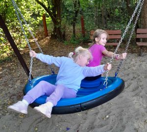 Dwie uczennice oddziału przedszkolnego bujają się na dużej niebieskiej huśtawce, której siedzisko jest w kształcie koła.