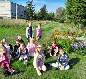 Uczniowie oddziału przedszkolnego siedzą na trawniku. Dwie dziewczynki z tyłu stoją. W tle widać blok.