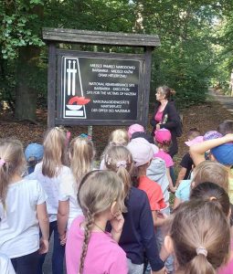 Pani nauczycielka stoi przy tablicy z napisem" "MIEJSCE PAMIĘCI NARODOWEJ BARBARKA - MIEJSCE KAŹNI OFIAR HITLERYZMU", "NATIONAL REMEMBERENCE SITE BARBARKA - EXECUTION SITE FOR THE VICTIMS OF NAZISM", "NATIONALGEDENKSTATE MARTEPLATZ DER OPFER DES NAZIMUS". Pani nauczycielka patrzy na tablicę. Na przeciwko stoją uczniowie oddziału przedszkolnego.