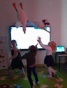 Troje dzieci z oddziału przedszkolnego bawi się na dywanie. Dzieci podrzucają w górę pluszowe gęsi.