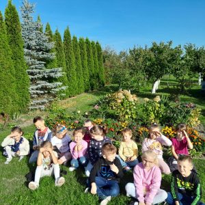 Uczniowie oddziału przedszkolnego siedzą na trawie przed ogródkiem działkowym. dzieci zasłaniają dłońmi oczy przed słońcem. W tle widać zielone drzewa owocowe i choinki równo przycięte i ustawione w rzędzie.