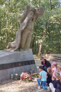 Zdjęcie pomnika ofiar zbrodni hitlerowskiej, przed którym klęczą uczniowie oddziału przedszkolnego wraz z nauczycielką. Pomnik jest w kształcie klęczącej postaci z ramionami złożonymi za plecami, spuszczoną głową i raną w piersi.