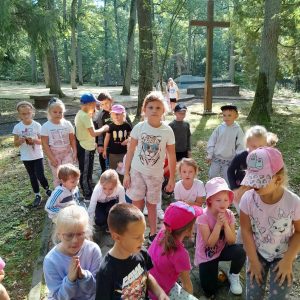 Uczniowie oddziału przedszkolnego stoją zgromadzeni na trawniku. Część z nich klęczy. Za nimi widać krzyż i pomnik ofiar ustawionych na Barbarce.