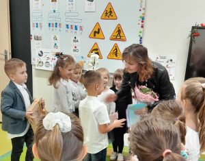 Przed wychowawczynią oddziału przedszkolnego stoi grupka uczniów. Pani pochyla się w kierunku jednego z uczniów. W dłoniach trzyma kwiaty i laurki.