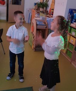 Dwoje uczniów oddziału przedszkolnego stoi obok siebie. Dzieci ubrane są w stroje galowe i patrzą się na siebie. Na dzieciach i podłodze odbijają się kolorowe światełka.