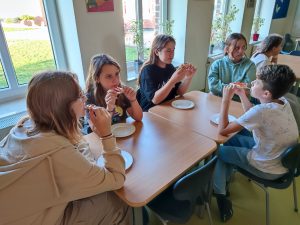 Uczniowie klasy szóstej siedzą przy stolikach na stołówce. Przed nimi leżą talerze. dzieci jedzą tosty.