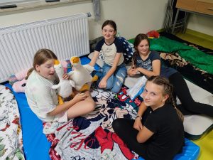 Uczniowie klasy szóstej siedzą na materacach na podłodze. Cztery dziewczynki na zdjęciu się uśmiechają. Jedna z nich przytula pluszową gęś.