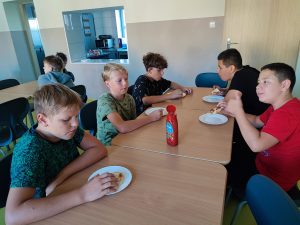 Uczniowie klasy szóstej siedzą przy stole na szkolnej stołówce. Chłopcy jedzą tosty. przed nimi stoją na stole talerze i ketchup.