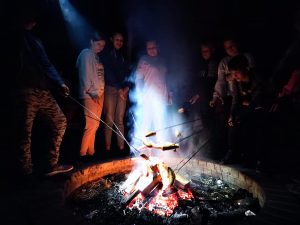 Uczniowie klasy szóstej stoją przy ognisku i smażą kiełbaski. Zdjęcie wykonane nocą.