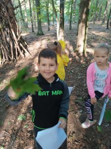 Na pierwszym planie widać uśmiechniętego chłopca trzymającego w wyciągniętej dłoni zielone liście oraz papierową torbę. W tle widać siwe dziewczynki.