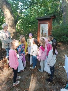 Uczniowie stoją przy obrazku oprawionym w drewniana ramę. Obok stoi Pani, na która dzieci patrzą.