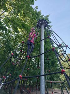 Uczennica wspięła się na szczyt siatki zamocowanej na metalowym słupie, na placu zabaw. Dziewczynka spogląda w dół.