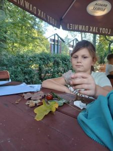 Uczennica siedzi przy dużej drewnianej ławie i spogląda na zebrane przez siebie dary jesieni - liście, żołędzie i kasztany.