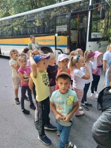 Uczniowie stoją w grupce przed autobusem. Jest z chłopców patrzy wprost w obiektyw. Reszta dzieci stoi bokiem.